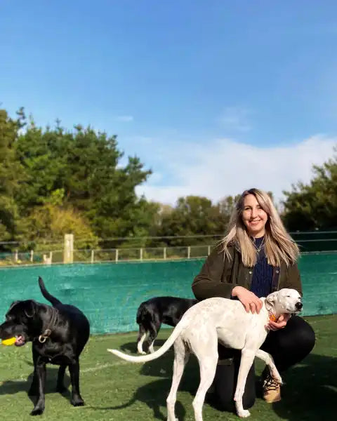 Katy Peck natural pet shop owner, sitting with a white dog in an outdoor setting with other dogs and greenery