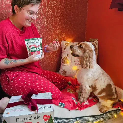 Person in red pajama set holding a Forthglade product next to a dog on a bed.