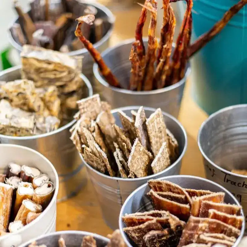 Metallic silver buckets filled with various types of dried pet treats, including jerky strips and bone-shaped snacks.