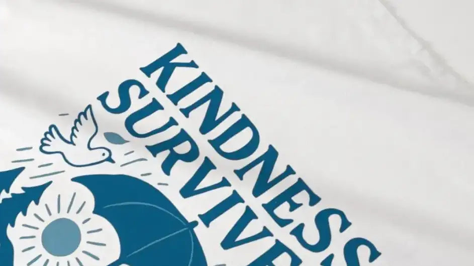 White t-shirt featuring a blue graphic with the text ’kindness survives’ and illustrations of a dove, an umbrella, and sunflowers.